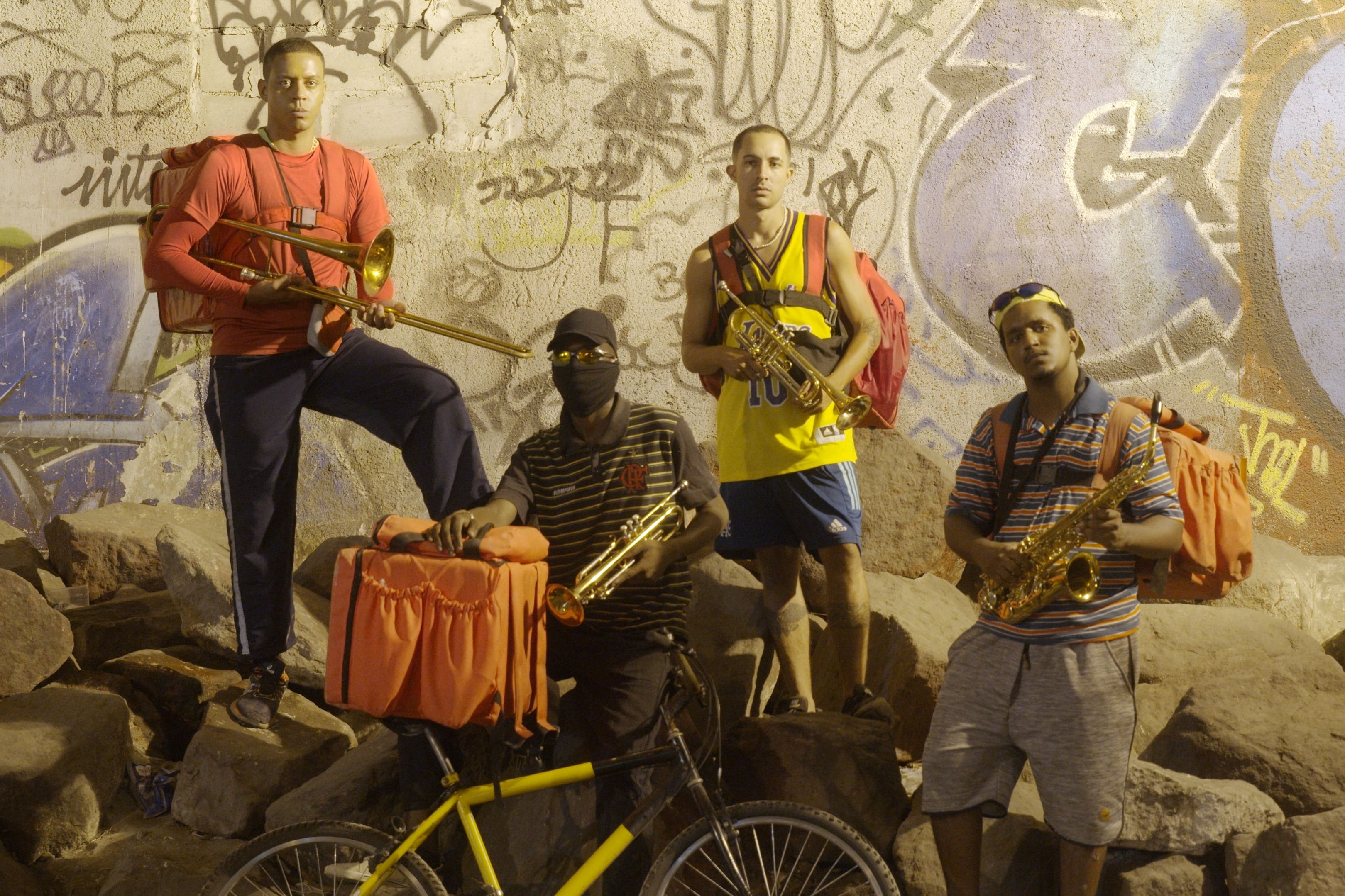 Quatro músicos de rua posam em frente a uma parede grafitada. Eles seguram trombone, trompete e saxofone, com mochilas nas costas. Um deles está com uma bicicleta amarela. A iluminação amarelada destaca os detalhes dos instrumentos e o cenário urbano ao redor.